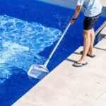 Person skims debris from a bright blue swimming pool using a long-handled net beside a metal ladder on sunny concrete.