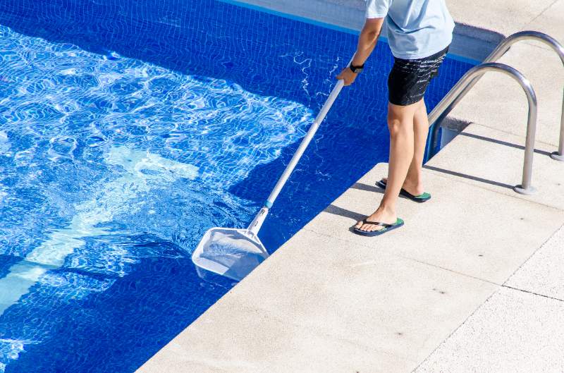 Person skims debris from a bright blue swimming pool using a long-handled net beside a metal ladder on sunny concrete.