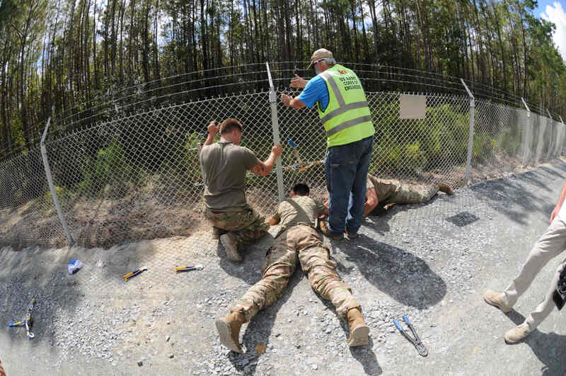 Several people are working to repair a damaged post of a chain-link fence along a roadside.