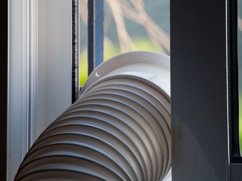 the exhaust process of a portable air conditioner, with a white pipe leading from the unit and out through a window using a white plastic vent kit.