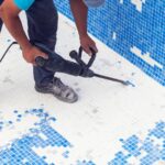 Worker uses a jackhammer to remove old blue mosaic pool tiles during resurfacing and renovation.