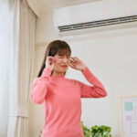 A woman stands in a room with an air conditioner on the wall, holding her hands to her ears and grimacing as if bothered by noise