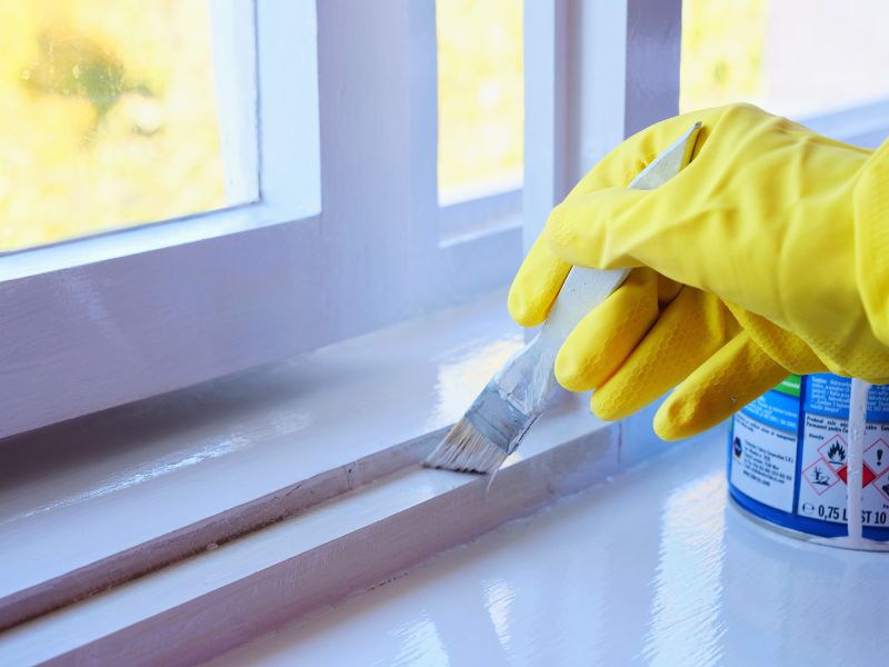 Hand in yellow rubber gloves applies a satin paint finish to window frame. 