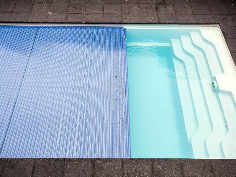 An automatic, corrugated blue pool cover is partially rolled up, revealing the light blue water and white steps of an outdoor in-ground swimming pool. The pool deck is made of dark gray pavers.