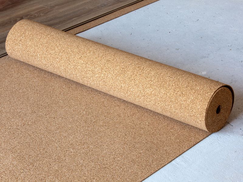 A roll of cork underlayment is unrolled on a concrete subfloor, with laminate planks visible nearby, showing its use for soundproofing.