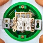 A close-up, top-down view of a double light switch being installed in a green electrical box. Exposed wires are visible on the terminal screws.