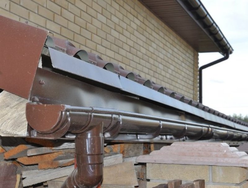Brown half-round gutter system with matching downspout installed on brick house, providing efficient rainwater drainage and durable roof protection.