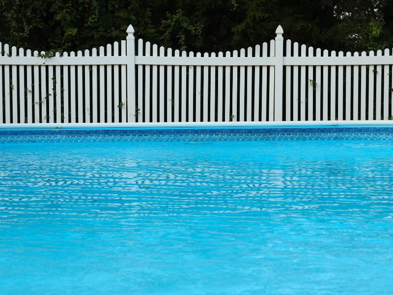 Swimming pool and white vinyl picket fence in front of trees 
