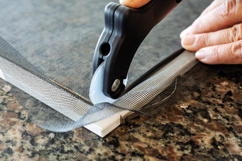 Close-up of hands using a spline roller tool to press mesh screen into window frame groove on a granite surface.