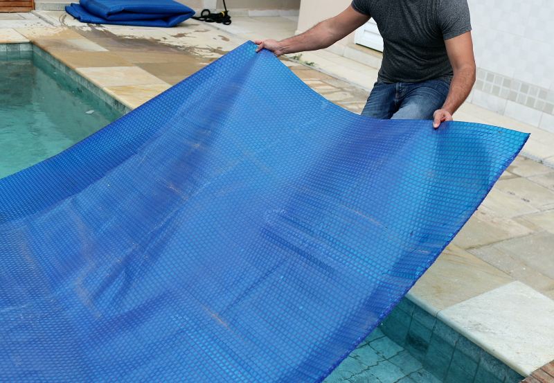 A man places a large blue bubble solar pool cover on a residential swimming pool, preparing for protection, heat retention, and reduced water evaporation.