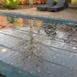 A dark, wet mesh pool cover is stretched over a rectangular pool, with pooled water and scattered autumn leaves sitting on the surface.