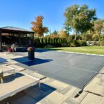 How to Install a Pool Cover on Your In-Ground Pool Backyard swimming pool covered with gray tensioned safety cover, surrounded by lounge chairs, patio, trees, and gazebo under clear blue sky.