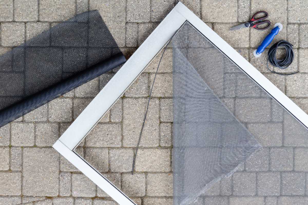 Screen door frame being repaired with new mesh, roll of screen material, spline, spline roller tool, and scissors on stone pavement.