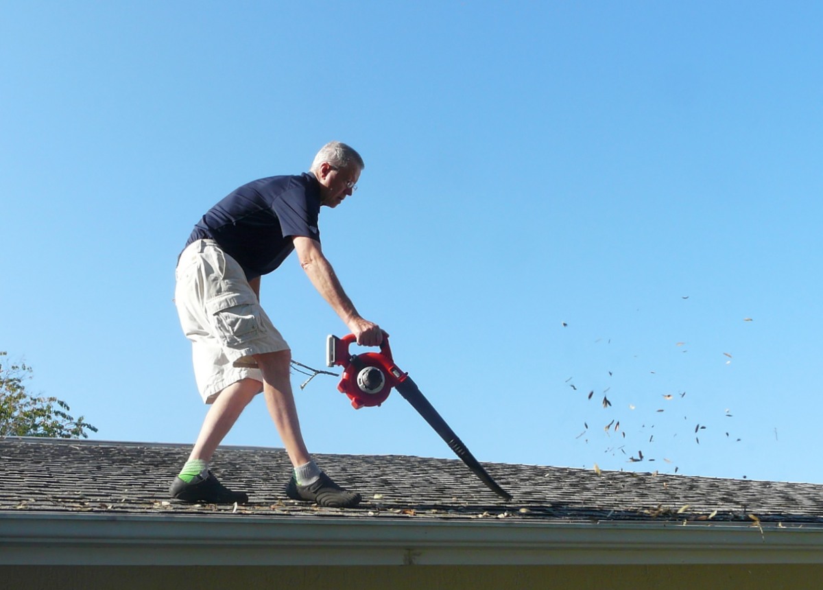 A homeowner is on the roof of his residence using a leaf blower to remove leaves from the gutter in preparation for storm season water runoff.