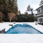Snow-covered backyard pool at residential home in winter, with partially frozen water, surrounding trees, and overcast sky creating a realistic scene