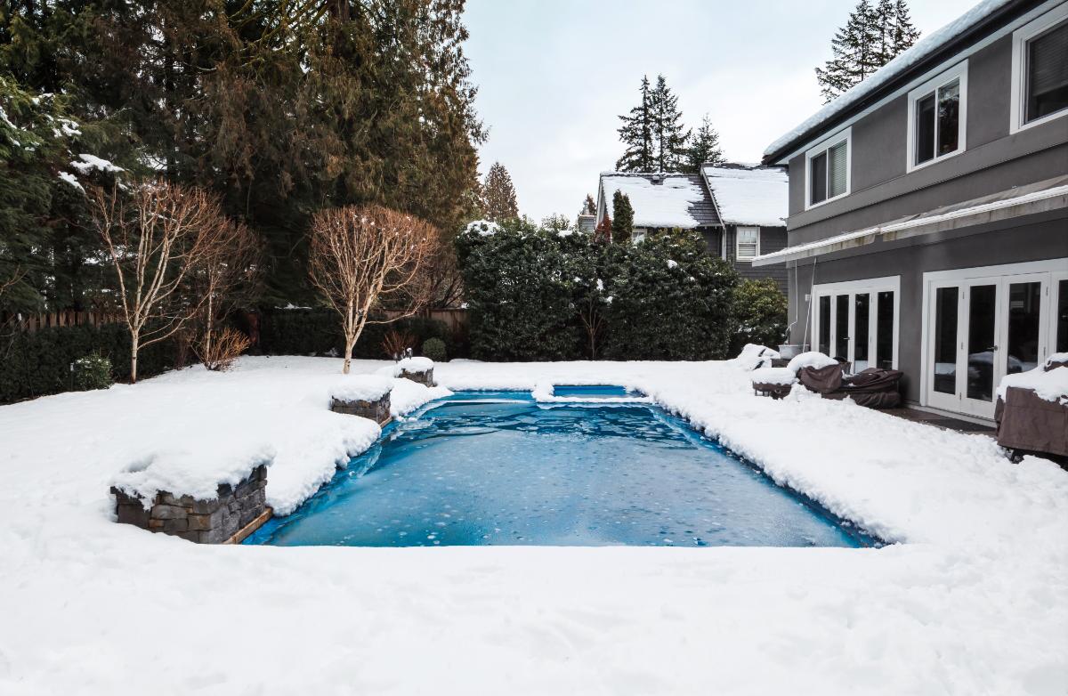 Snow-covered backyard pool at residential home in winter, with partially frozen water, surrounding trees, and overcast sky creating a realistic scene