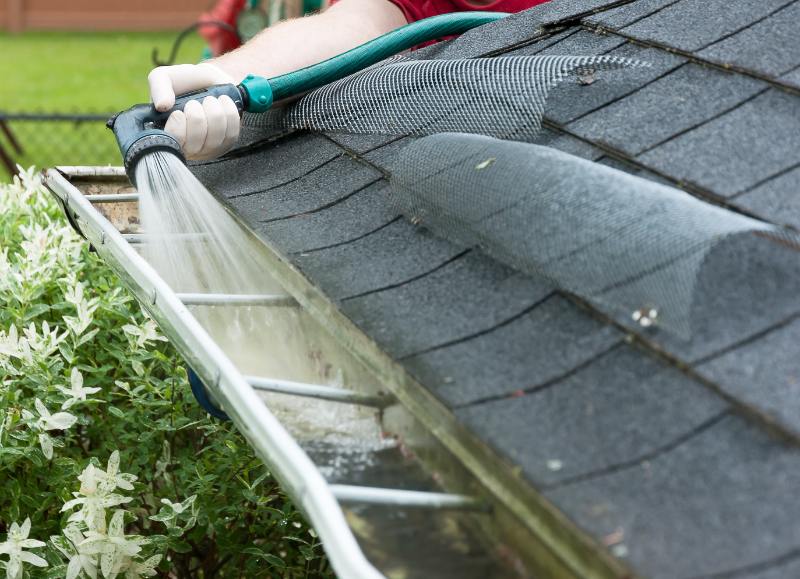 Man standing on a ladder cleaning house gutters with a garden hose, washing away dirt and debris from the roof edge.