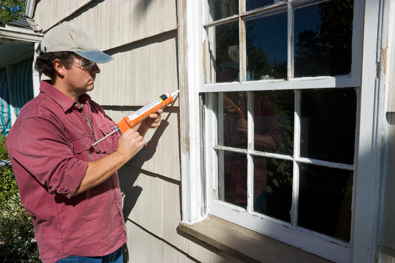 Applying caulk with a caulk gun around an old wooden window frame, demonstrating caulking between the window and the siding.