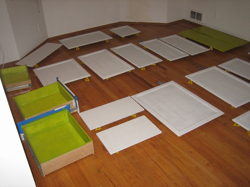 Numerous cabinet doors and drawer faces are laid out on a wood floor and elevated by stands, showing them being primed white and painted green.