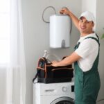 Plumber in green overalls replacing wall mounted boiler, standing beside washing machine with tool bag in bright modern laundry room.