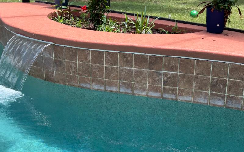 White calcium and mineral deposits visible along the waterline tile and grout lines of a residential pool with a spillway water feature, turquoise water, and coral-colored pool coping.